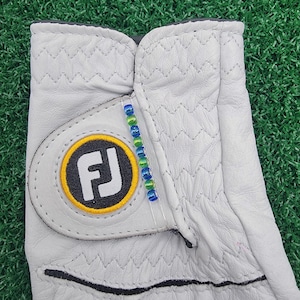 May include: A light gray golf glove with a stitched diamond pattern. The glove features a circular logo with the letters "FJ" in black and white, surrounded by a yellow circle. A beaded accent in blue and green is attached to the glove.