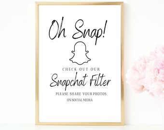 以下が含まれることがあります： Snapchatのゴーストロゴと「Oh Snap!」というテキストが書かれた、金色のフレームが付いた白い看板。看板には、「Check out our Snapchat Filter」と「Please share your photos on social media.」というテキストも書かれています。