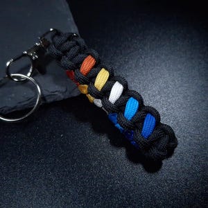 May include: A keychain featuring a braided paracord fob in a black exterior weave with a multi-colored interior core showing red, orange, yellow, white, light blue, and dark blue segments, attached to a silver-toned metal lobster clasp and ring.
