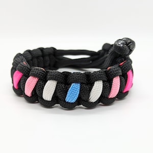 May include: A black paracord bracelet with pink, white, and blue stripes. The bracelet is tied with a knot at the end.