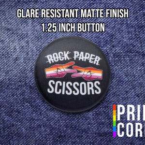 May include: A 1.25-inch black button with the text "Rock Paper Scissors" and a graphic of hands in the scissors and paper positions. The button has a matte finish and the Pride Cords logo is visible.