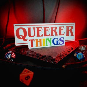 May include: A sticker with the text "QUEERER THINGS" in red and rainbow colors. Several polyhedral dice are scattered around the sticker, with a dark background and red lighting.