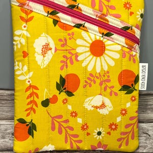 May include: A yellow quilted pouch with a floral and fruit design. The pattern features white daisies, orange fruits, and pink accents. The zip is bright pink, and the text "Stitch Chick Design" is visible on a small tag.