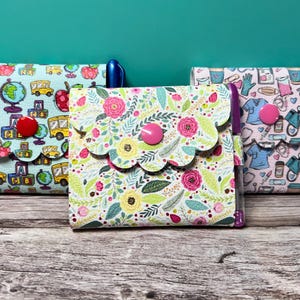 May include: Three fabric pouches with scalloped edges and button closures. One pouch features a school-themed print with apples, globes, and school buses. Another has a floral pattern, and the third displays a medical theme.