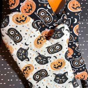 May include: A pair of Halloween-themed oven mitts. One mitt is black with a white star pattern and features images of black cats, owls, and jack-o'-lanterns. The other mitt is white with a black star pattern and features images of black cats, owls, and jack-o'-lanterns.
