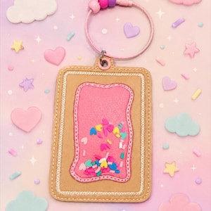 May include: A rectangular keychain with a pink interior filled with colorful confetti. The keychain has a tan border with pink stitching and a pink wristlet with pink and purple beads. Decorative hearts, stars, and clouds surround the keychain.