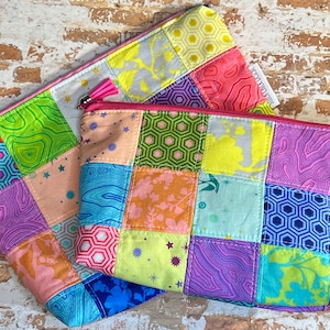 Pode incluir: Duas bolsas de zíper de patchwork coloridas com borlas rosa. As bolsas são feitas de diferentes tecidos com padrões geométricos e florais em tons de rosa, azul, verde, amarelo e roxo.