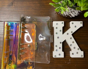 May include: A clear plastic binder with a holographic cover and a clear plastic pocket on the front. The binder has six rings and a silver button closure. A white wooden letter K with rhinestones is in the background.