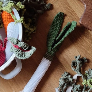 May include: Crocheted vegetables including a leek, carrot, and leafy greens. The leek has a white stem and green leaves. The carrot is orange, and the greens are various shades of green. The items are arranged on a wooden surface.