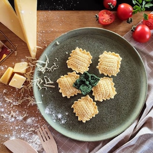 May include: A plate of crocheted ravioli with a green crocheted basil leaf on top. The plate is green and has a white rim. There is grated cheese and a wooden spoon on the table.