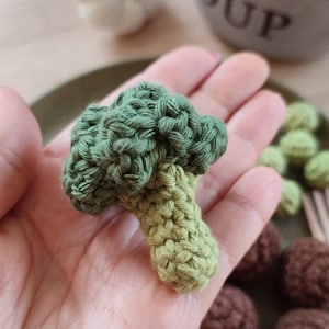 May include: A crocheted green broccoli with a light green stem. The broccoli is being held in a hand. There are brown crocheted balls in the background.