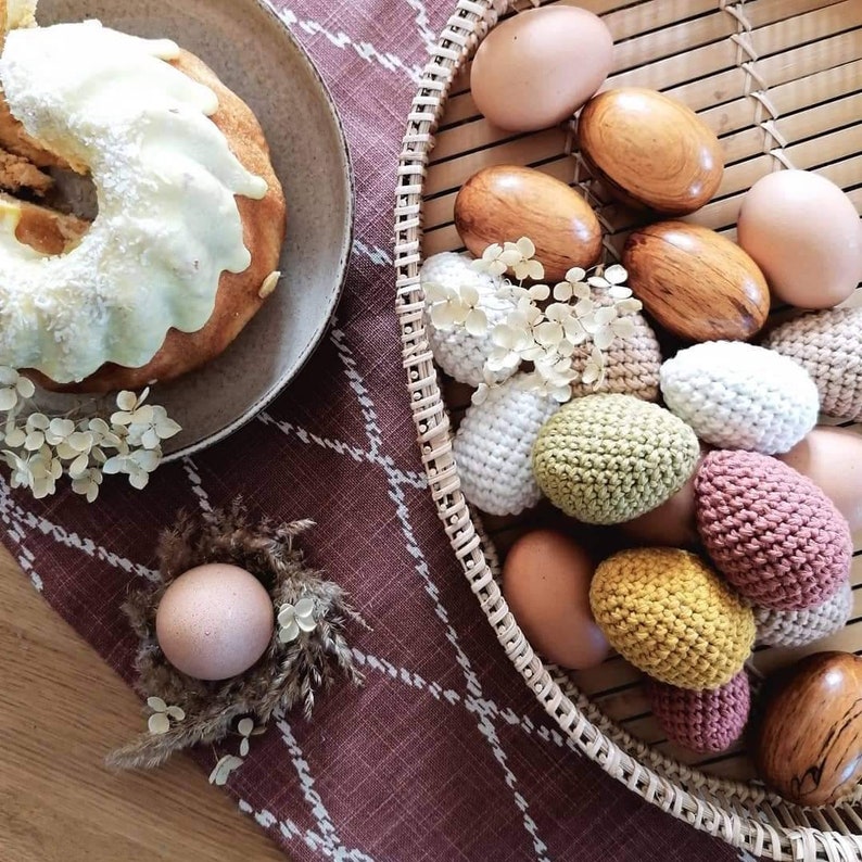 Pu&ograve; includere: Un cesto di vimini pieno di uova di Pasqua all'uncinetto e di legno. Le uova sono in tonalit&agrave; di marrone, bianco, verde e rosa. C'&egrave; anche una torta bianca con glassa bianca sullo sfondo.