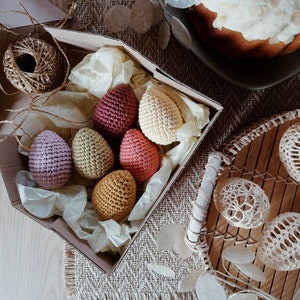 Pu&ograve; includere: Una scatola piena di uova di Pasqua all'uncinetto in vari colori, tra cui rosa, rosso, giallo e beige. Le uova sono disposte in una scatola con carta velina e sono circondate da una scatola di cartone marrone.