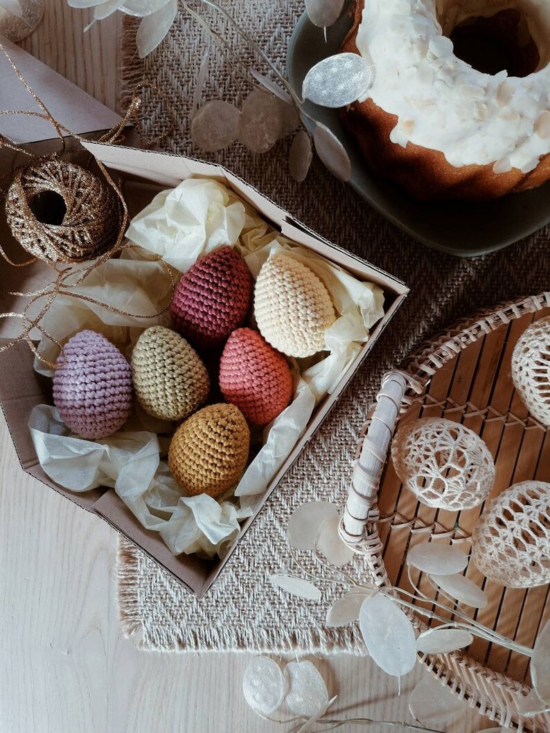 Pu&ograve; includere: Una scatola piena di uova di Pasqua all'uncinetto in vari colori, tra cui rosa, rosso, giallo e beige. Le uova sono disposte in una scatola con carta velina e sono circondate da una scatola di cartone marrone.