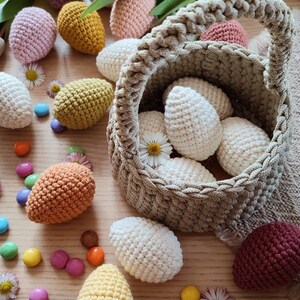 Pu&ograve; includere: Un cesto all'uncinetto pieno di uova all'uncinetto colorate. Il cesto &egrave; fatto di un filo beige e ha un manico intrecciato. Le uova sono di vari colori, tra cui bianco, rosa, giallo, arancione e viola. Le uova sono sparse intorno al cesto su una superficie di legno.