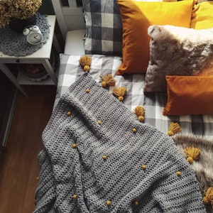 Op de afbeelding: Een grijze gehaakte deken met gele pompoenen en grijs-wit geruit beddengoed. De deken heeft een textuurpatroon en is versierd met kleine gele pompoenen.