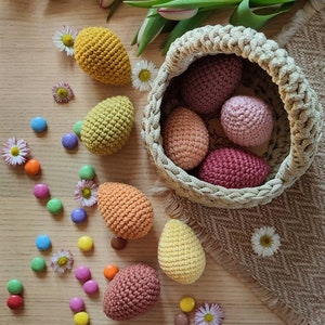 Pu&ograve; includere: Un cestino di Pasqua all'uncinetto pieno di uova all'uncinetto colorate. Il cestino &egrave; fatto di filato beige e si trova su una superficie di legno. Ci sono anche alcuni dolci colorati sparsi intorno al cestino.