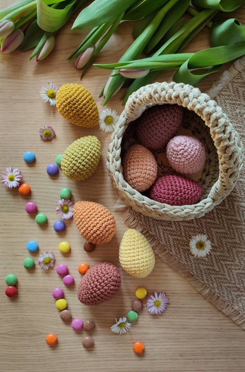 Pu&ograve; includere: Un cestino di Pasqua all'uncinetto pieno di uova all'uncinetto colorate. Il cestino &egrave; fatto di filato beige e si trova su una superficie di legno. Ci sono anche alcuni dolci colorati sparsi intorno al cestino.