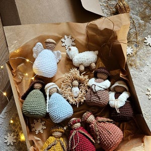 K&ouml;nnte beinhalten: Eine Sammlung geh&auml;kelter Weihnachtsschmuckst&uuml;cke in einer Pappschachtel. Das Set umfasst Engel, ein Lamm und Figuren in verschiedenen Farben wie Wei&szlig;, Blau, Gr&uuml;n, Gelb und Rot. Die Schachtel ist mit braunem Papier ausgekleidet und mit Schneeflocken-Ornamenten verziert.