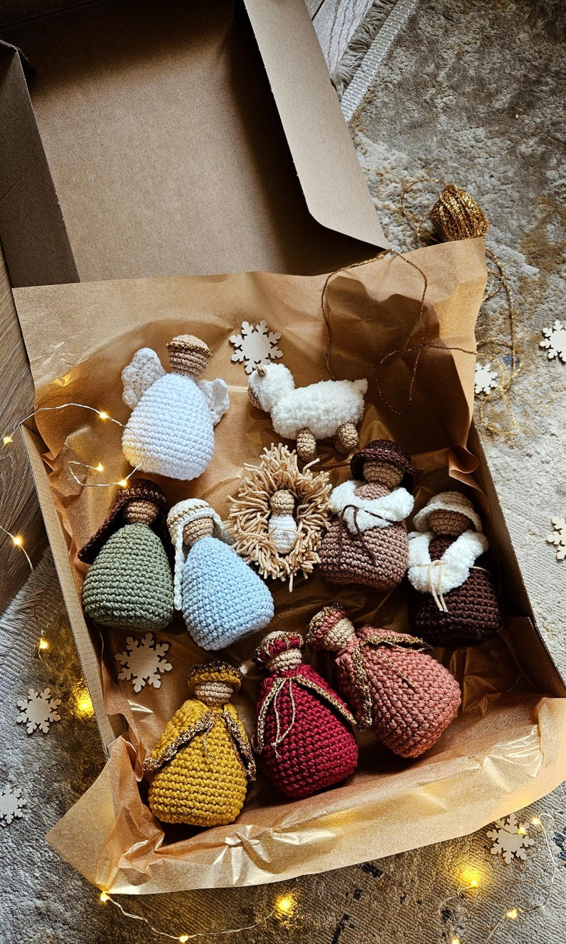 K&ouml;nnte beinhalten: Eine Sammlung geh&auml;kelter Weihnachtsschmuckst&uuml;cke in einer Pappschachtel. Das Set umfasst Engel, ein Lamm und Figuren in verschiedenen Farben wie Wei&szlig;, Blau, Gr&uuml;n, Gelb und Rot. Die Schachtel ist mit braunem Papier ausgekleidet und mit Schneeflocken-Ornamenten verziert.