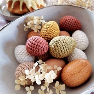 Pu&ograve; includere: Una ciotola piena di uova di Pasqua all'uncinetto in vari colori, tra cui beige, marrone, verde e rosa. Le uova sono circondate da fiori bianchi secchi.