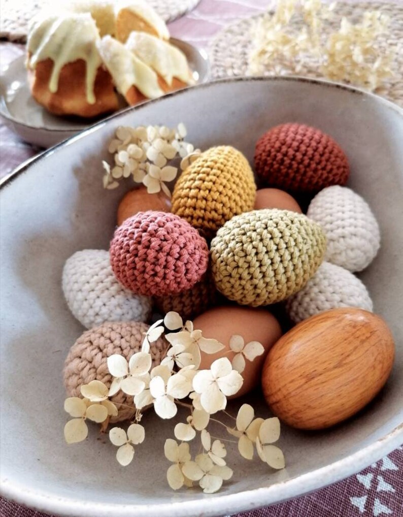 Pu&ograve; includere: Una ciotola piena di uova di Pasqua all'uncinetto in vari colori, tra cui beige, marrone, verde e rosa. Le uova sono circondate da fiori bianchi secchi.