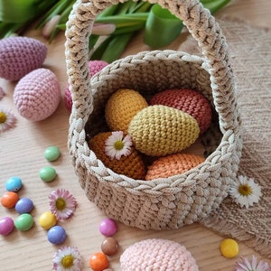Pu&ograve; includere: Un cestino all'uncinetto con manico, pieno di uova all'uncinetto colorate. Il cestino &egrave; fatto di filato beige e le uova sono in tonalit&agrave; di rosa, giallo, arancione e rosso. Ci sono anche alcuni dolci colorati sparsi intorno al cestino.