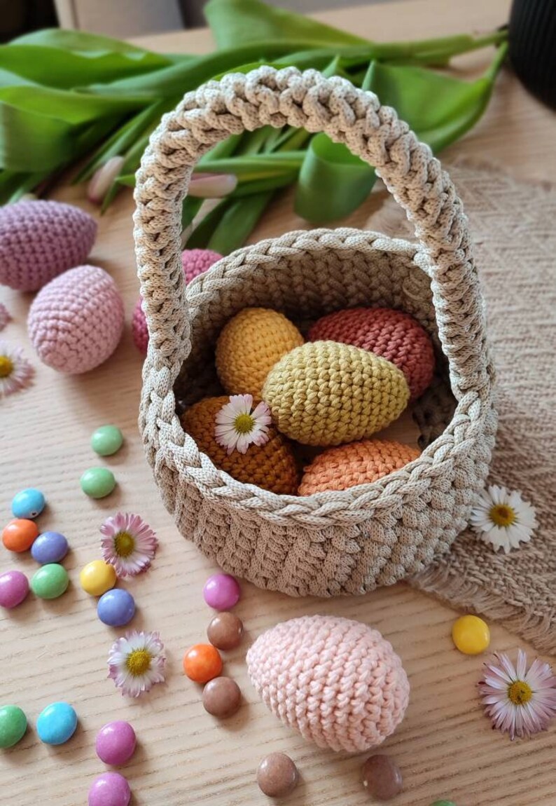 Pu&ograve; includere: Un cestino all'uncinetto con manico, pieno di uova all'uncinetto colorate. Il cestino &egrave; fatto di filato beige e le uova sono in tonalit&agrave; di rosa, giallo, arancione e rosso. Ci sono anche alcuni dolci colorati sparsi intorno al cestino.