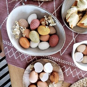 Pu&ograve; includere: Una ciotola bianca piena di uova di Pasqua all'uncinetto in varie tonalit&agrave; di marrone, giallo e rosa. La ciotola &egrave; su un tavolo con una tovaglia a motivi marroni e bianchi. Ci sono altre uova all'uncinetto su un piatto di legno e un piatto bianco con uova vere.
