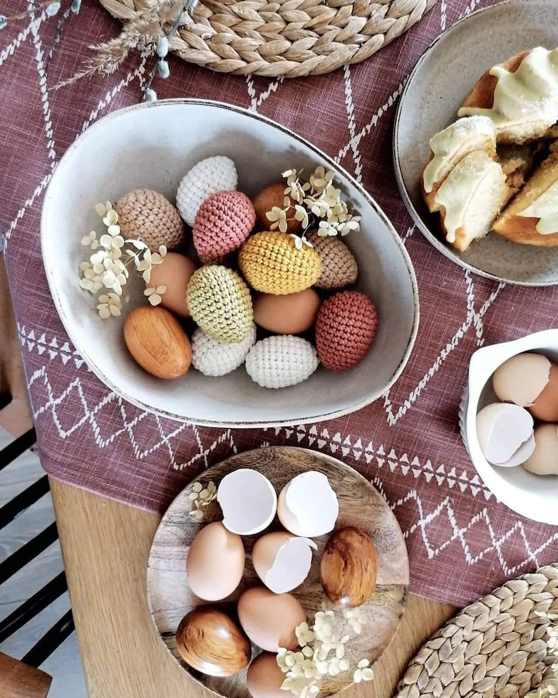 Pu&ograve; includere: Una ciotola bianca piena di uova di Pasqua all'uncinetto in varie tonalit&agrave; di marrone, giallo e rosa. La ciotola &egrave; su un tavolo con una tovaglia a motivi marroni e bianchi. Ci sono altre uova all'uncinetto su un piatto di legno e un piatto bianco con uova vere.