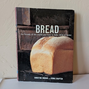May include: A hardcover book titled BREAD: the breads of the world and how to bake them at home by Christine Ingram and Jennie Shapter. The cover features a large, golden-brown loaf of bread resting on a dark metal rack against a dark background.