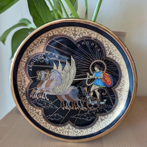May include: A round decorative plate with a black center and gold rim. The design features a figure in a blue tunic driving a chariot pulled by three winged horses. The text APOLLON and RHODES is printed in gold. Intricate gold patterns border the edge.
