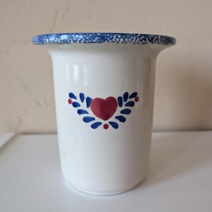May include: A white ceramic utensil holder with a flared rim featuring a blue speckled pattern. The front of the vessel is decorated with a hand-painted red heart centered between two symmetrical blue leaf motifs and small red dots.