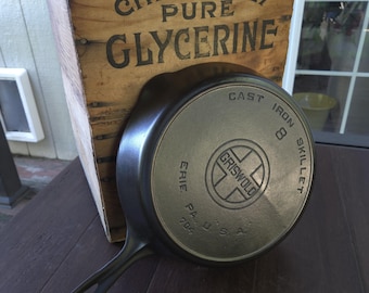 May include: A black cast iron skillet with a handle, featuring the embossed text 'GRISWOLD', 'CAST IRON', '8', 'SKILLET', 'ERIE PA. U.S.A.', and '704' on the bottom. It is positioned in front of a wooden crate labeled 'CHEMICALLY PURE GLYCERINE'.