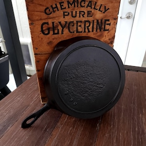 May include: A black cast iron skillet with a wooden handle. The skillet is sitting on a wooden table. A wooden crate with the text "Chemically Pure Glycerine" is behind the skillet.
