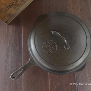 Pode incluir: Uma frigideira de ferro fundido cinza escuro com tampa e alça, repousando sobre uma superfície de madeira. A tampa apresenta um logotipo circular. Uma caixa de madeira é parcialmente visível no canto superior esquerdo.
