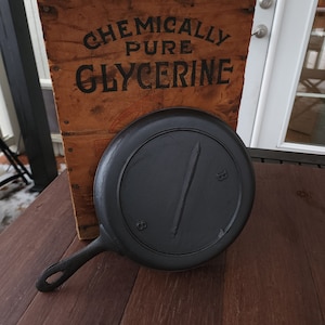 May include: A black cast iron skillet with a wooden handle. The skillet is sitting on a wooden surface. The text "CHEMICALLY PURE GLYCERINE" is visible on a wooden sign behind the skillet.