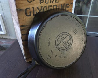 May include: A black cast iron skillet with a handle, featuring the embossed text 'GRISWOLD', 'ERIE PA., U.S.A.', and '10' on the underside. The skillet is positioned against a wooden crate labeled 'CHEMICALLY PURE GLYCERINE' in black lettering.