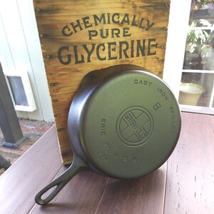 以下が含まれることがあります： 「Griswold」と「Cast Iron Skillet」の文字が底に書かれた鋳鉄製のフライパン。フライパンは、「Chemically Pure Glycerine」と書かれた木製の看板の前にある木製の表面に置かれています。