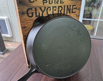 May include: A black cast iron skillet with the word ERIE and the number 9 stamped on the bottom, positioned in front of a wooden crate panel featuring the black text CHEMICALLY PURE GLYCERINE with visible metal nails along the top edge.