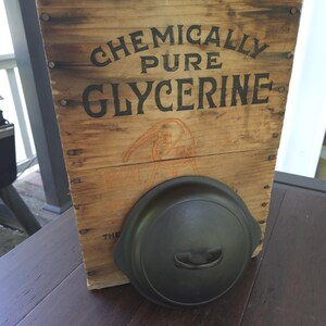 Può includere: Una cassa di legno con le parole "CHEMICALLY PURE GLYCERINE" stampate in nero. Un coperchio in ghisa nero poggia davanti alla cassa. La cassa è in legno e ha un aspetto invecchiato.