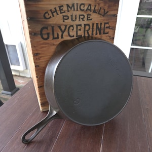 May include: A black cast iron skillet with a long handle, resting on a wooden surface. The skillet is positioned in front of a wooden crate with the text "CHEMICALLY PURE GLYCERINE" printed on it. The skillet has the number "9" on it.