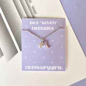 May include: A silver necklace with a heart charm and the number seven pendant, displayed on a light purple card. The card has the text "BTS 'seven' necklace" and "21centurygirls" printed on it, with small heart illustrations.