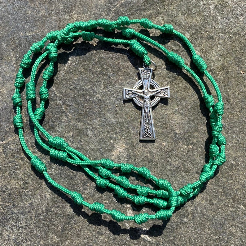 May include: A green knotted rosary with a silver Celtic cross pendant. The rosary is made of braided cord with knots at regular intervals.