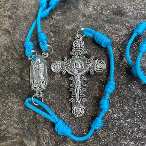 May include: A turquoise rosary with a silver crucifix and a pendant depicting a religious figure. The crucifix is adorned with rose details. The rosary beads are knotted, and the background is a textured gray surface.