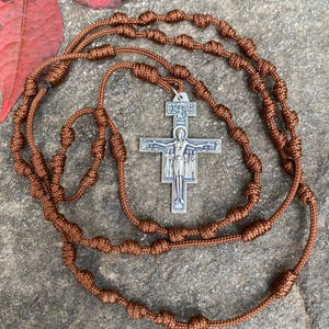 May include: A brown knotted rosary with a silver crucifix. The crucifix features a detailed depiction of Jesus Christ. The rosary beads are evenly spaced along the cord. The background is a textured, neutral surface.