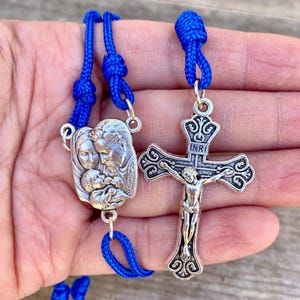 May include: A blue cord rosary with a silver crucifix and a silver pendant depicting the Holy Family. The pendant features a depiction of Mary, Joseph, and baby Jesus.