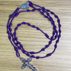 May include: A purple cord rosary featuring a silver-toned metal crucifix with a floral rose design and a central silver-toned medal depicting a religious figure. The rosary is constructed with knotted cord segments serving as beads in a continuous loop.