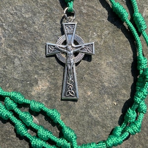 May include: A green knotted rope rosary with a silver Celtic cross pendant featuring a depiction of Jesus Christ. The cross has a circular halo with the inscription "INRI" in the center.