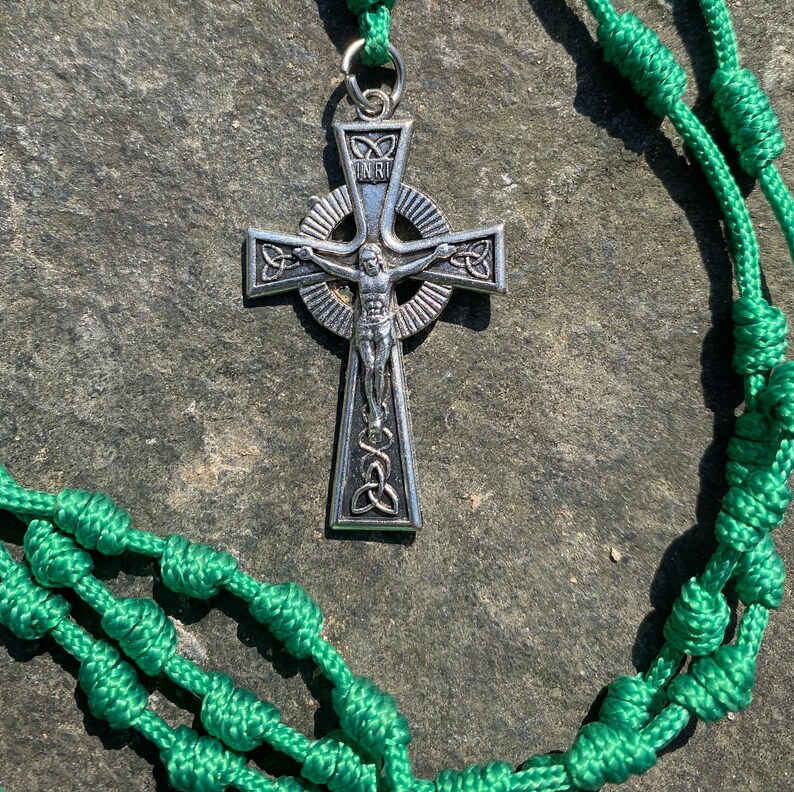 May include: A green knotted rope rosary with a silver Celtic cross pendant featuring a depiction of Jesus Christ. The cross has a circular halo with the inscription "INRI" in the center.